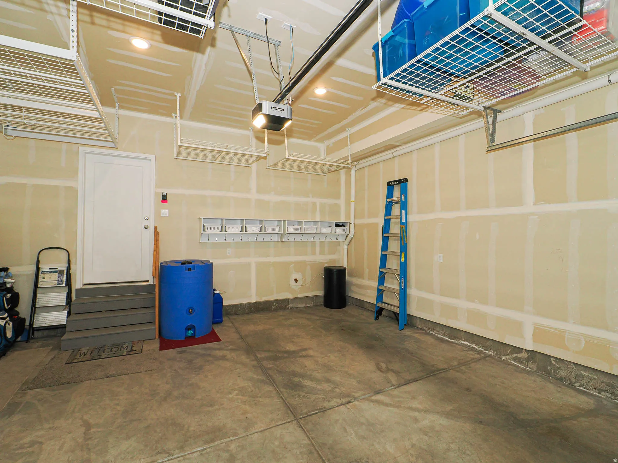 Spacious garage featuring a concrete floor, overhead storage racks, a white access door with steps, and exposed drywall
