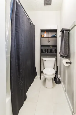 Bathroom featuring a standard toilet, light-colored tile flooring, and a shower/tub combination with a dark shower curtain