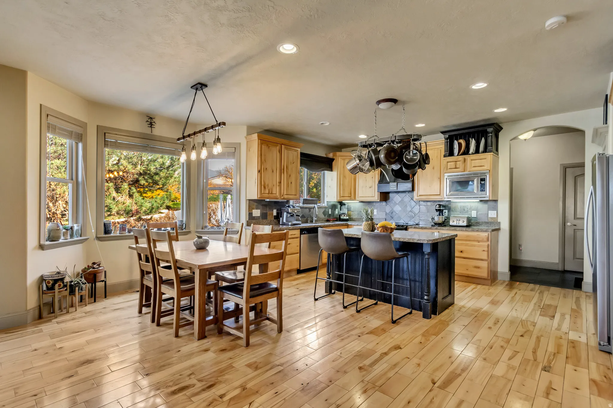Open-concept kitchen and dining area featuring light wood-finish flooring, a central island with a dark base and seating, light wood cabinetry, stainless steel appliances, and multiple windows