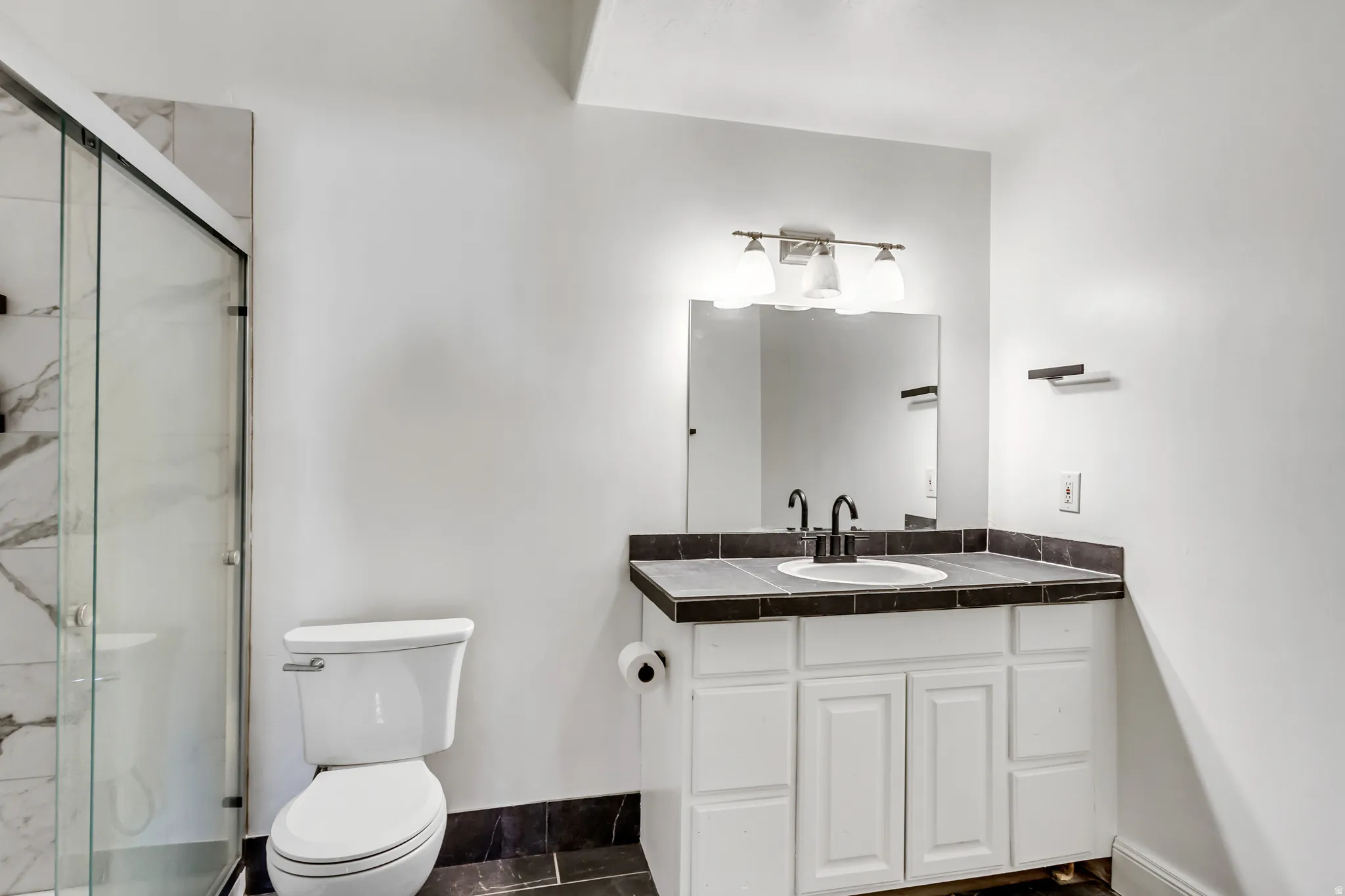 Bathroom featuring a glass-enclosed shower with marble-look tile, white vanity with a dark tiled countertop and integrated sink, black fixtures, and a wall-mounted mirror with overhead lighting