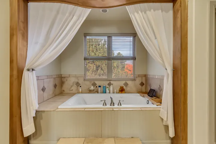 Built-in soaking tub with deck-mounted faucet, surrounded by tiled walls with diamond accents, and a window with horizontal blinds