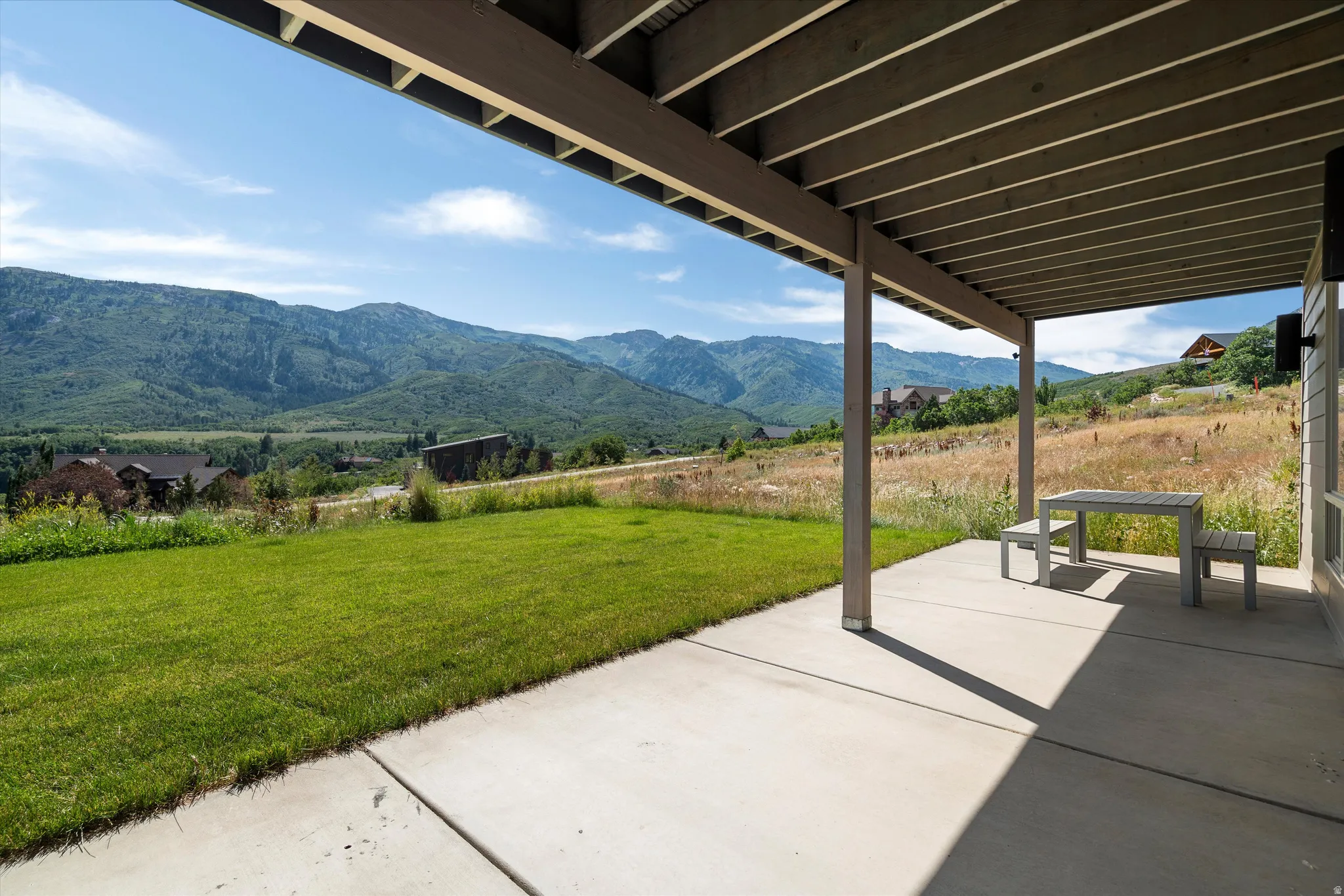 Covered outdoor patio with concrete flooring and exposed beam ceiling, opening onto a grassy lawn