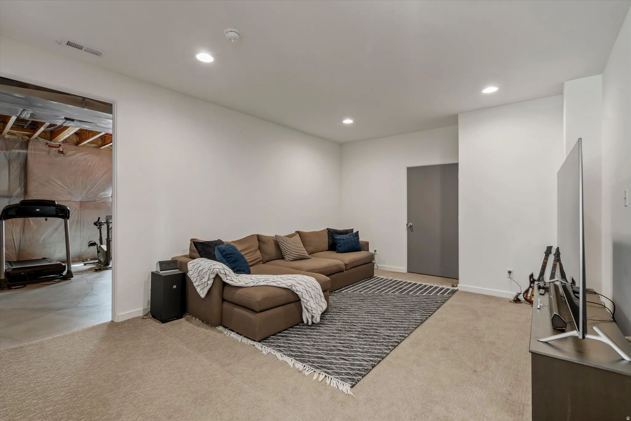 Recessed lighting illuminates this versatile room with carpet flooring and crisp white walls, featuring a solid gray door and an open doorway revealing an unfinished space