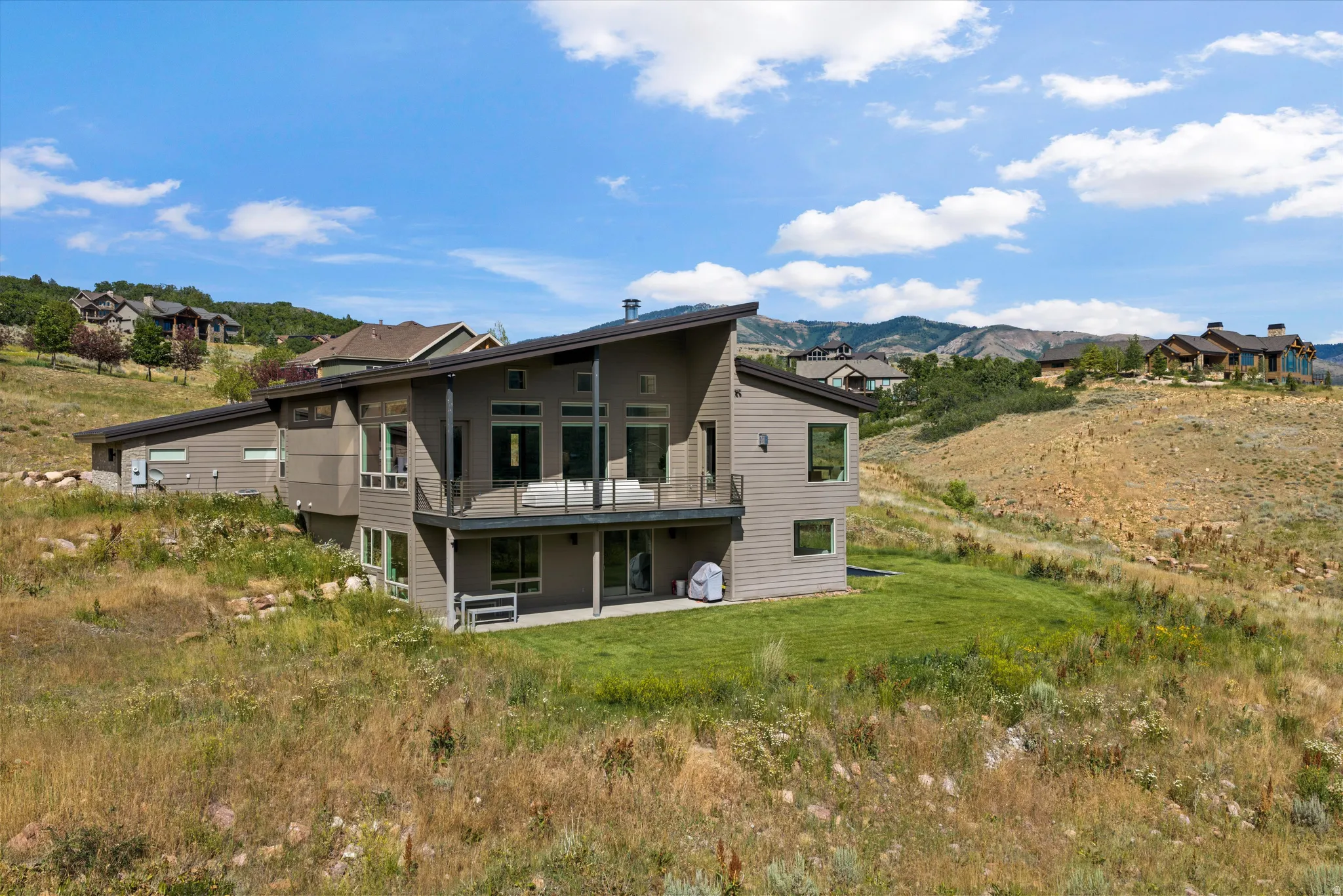Contemporary multi-level residence featuring a sloped roofline, a second-story balcony with glass railings, an outdoor patio, expansive windows, and a neutral-toned siding exterior