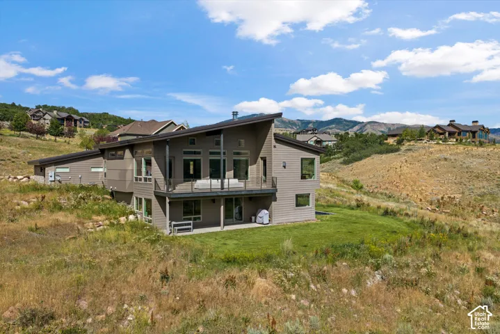 Contemporary multi-level residence featuring a sloped roofline, a second-story balcony with glass railings, an outdoor patio, expansive windows, and a neutral-toned siding exterior