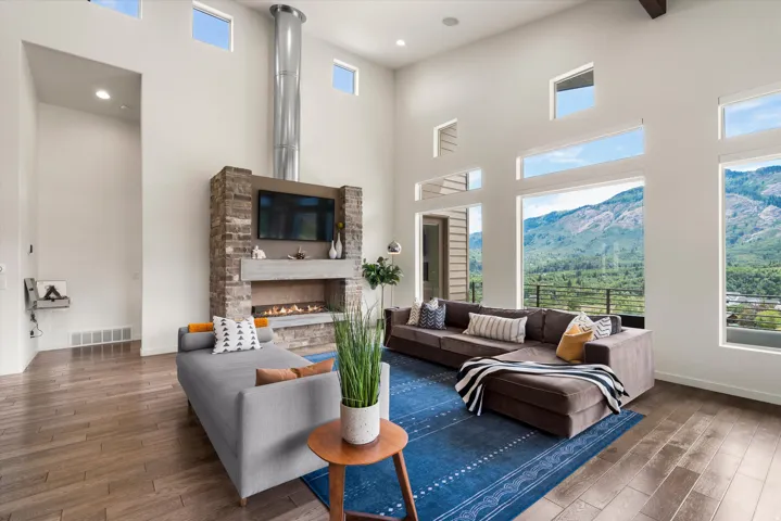 Spacious living area featuring high ceilings, wood-finish flooring, a stone-clad fireplace with a built-in television mount, and extensive window walls showcasing mountain scenery