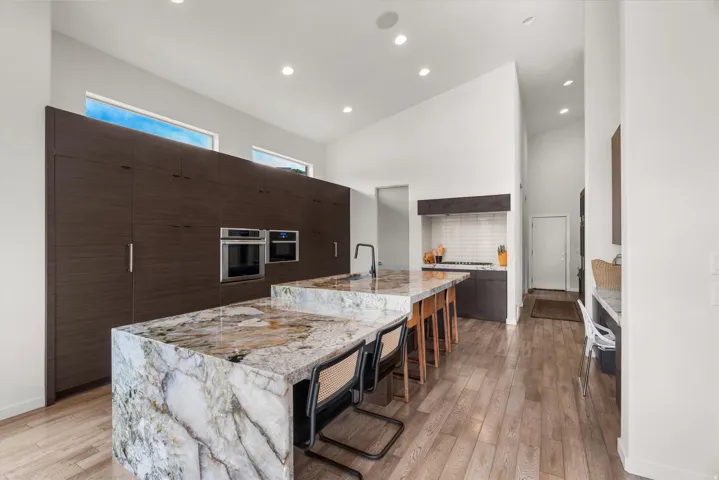 Contemporary kitchen featuring two large islands with stone countertops, integrated stainless steel appliances, extensive dark wood-finish cabinetry, light wood-finish flooring, and high vaulted ceilings with recessed lighting