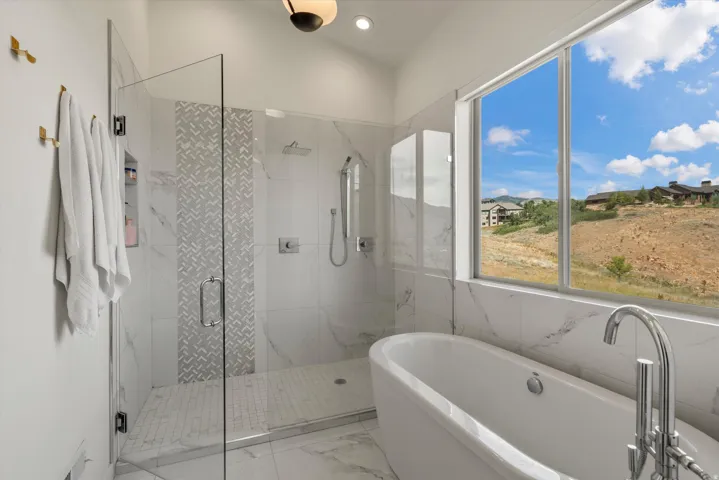 Bathroom featuring a freestanding soaking tub with floor-mounted chrome faucet, a large window, and a glass-enclosed shower with a rain shower head, hand-held shower, and patterned tile accent