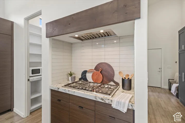 Built-in gas cooktop with a stainless steel range hood, stone countertop, wood-finish cabinetry, and a walk-in pantry with built-in shelving