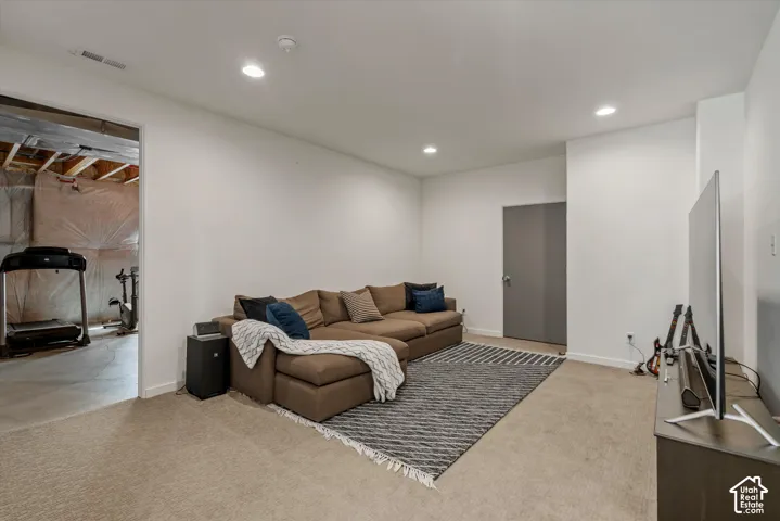 Recessed lighting illuminates this versatile room with carpet flooring and crisp white walls, featuring a solid gray door and an open doorway revealing an unfinished space