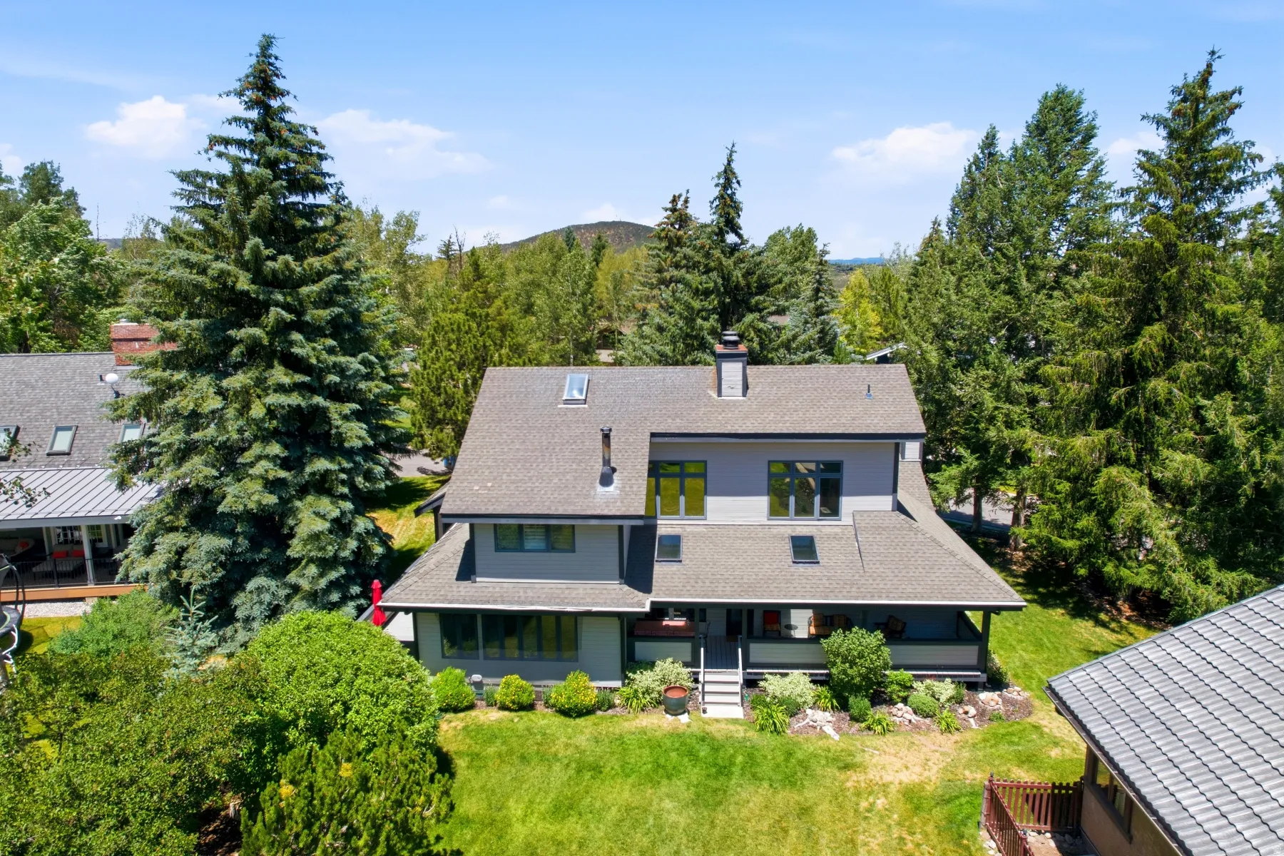 Multi-level architecture with a gray exterior, a shingled roof with multiple skylights, a covered porch, and a landscaped front yard with grass and mature trees