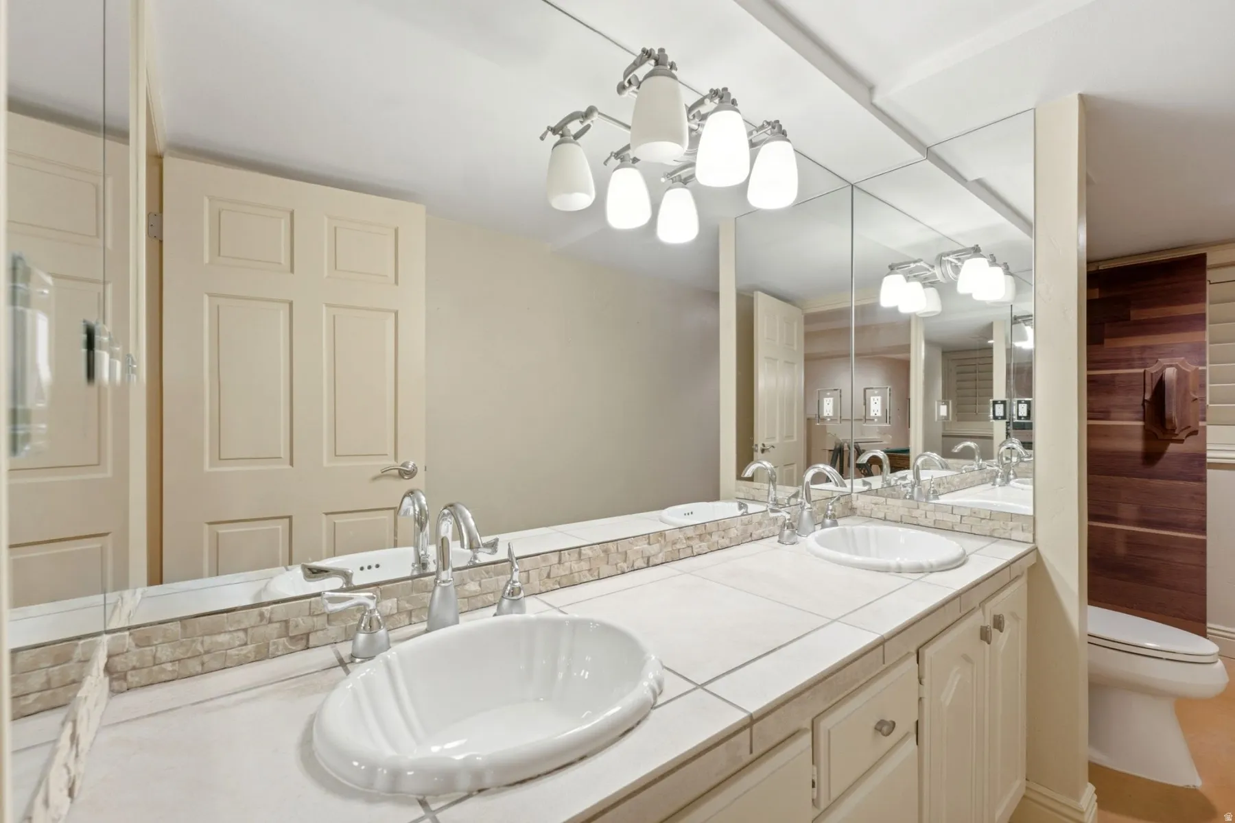 Dual oval undermount sinks, tile countertop, integrated vanity cabinetry, full-width mirror, and wall-mounted light fixture