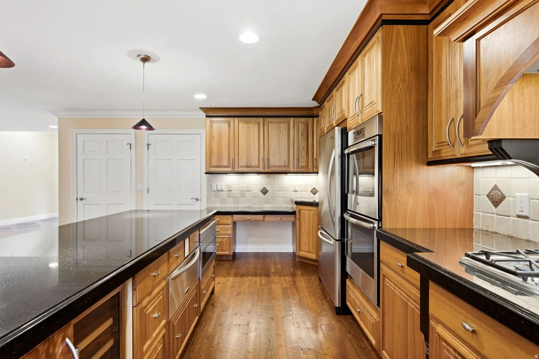 Custom wood cabinetry with crown molding, black stone countertops, stainless steel double ovens, stainless steel refrigerator, and wood-finish flooring