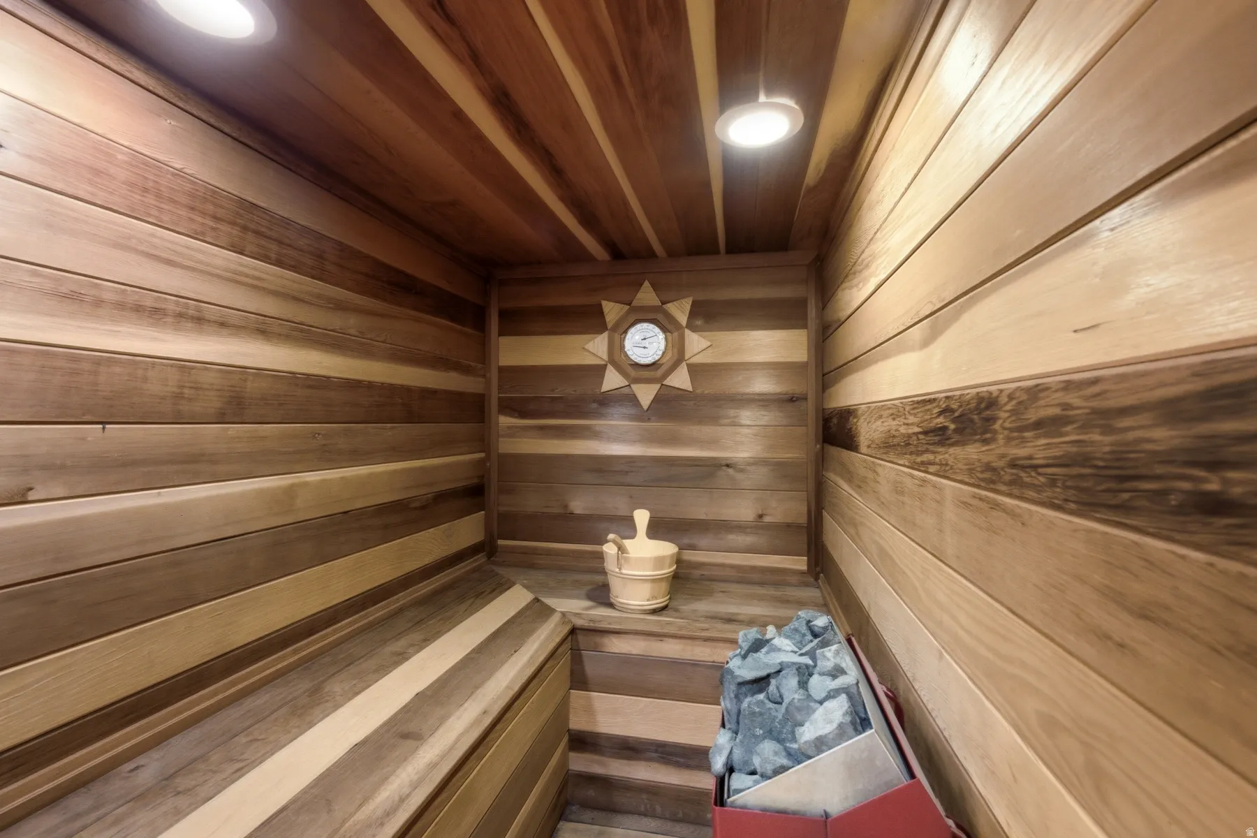 Wood-paneled sauna featuring built-in benches, recessed ceiling lighting, and a circular thermometer with decorative wood surround