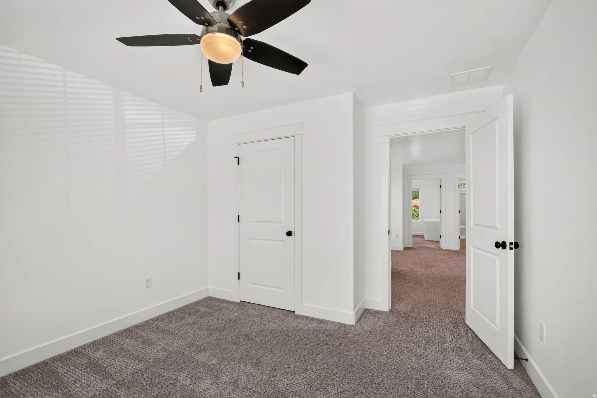Spacious room featuring neutral carpeting, white trim, a multi-blade ceiling fan, and a solid core door with black hardware
