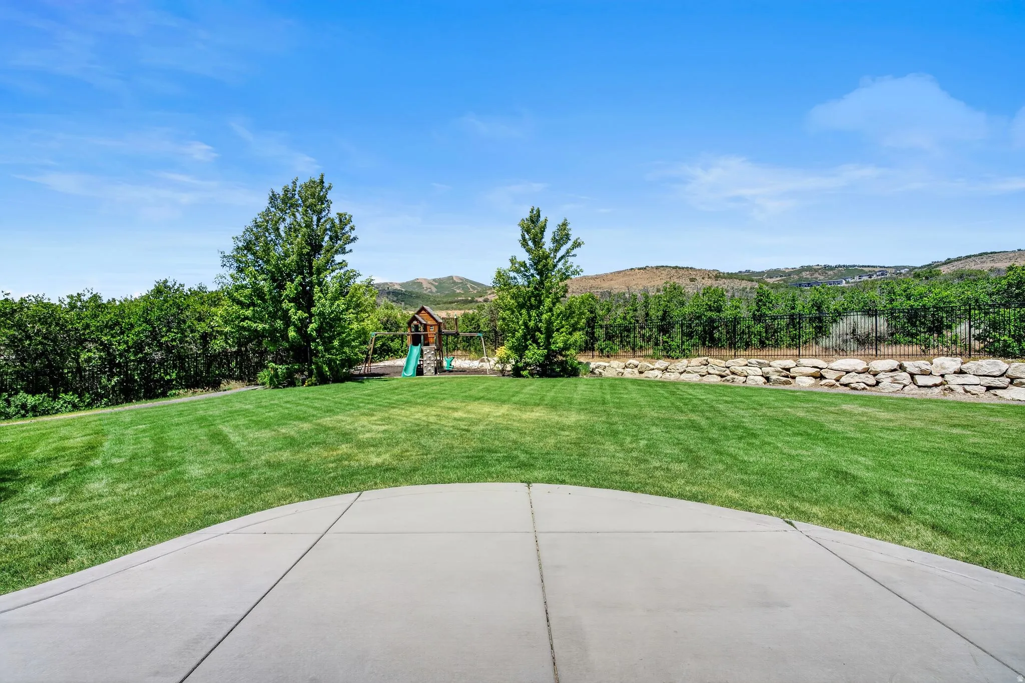 Expansive backyard featuring a large concrete patio, lush green lawn, mature trees, and a rock retaining wall