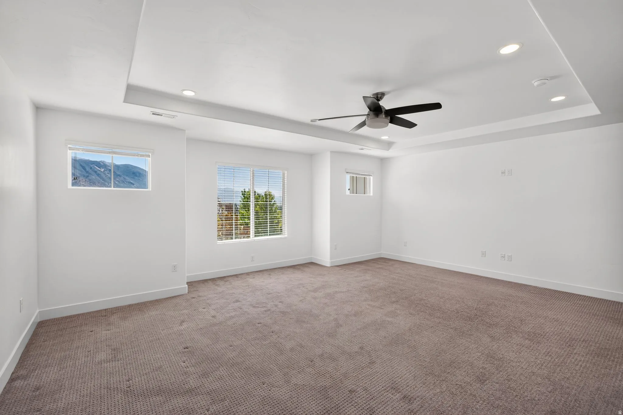 Spacious interior room featuring a tray ceiling with recessed lighting and a ceiling fan, plush carpet flooring, and white baseboards
