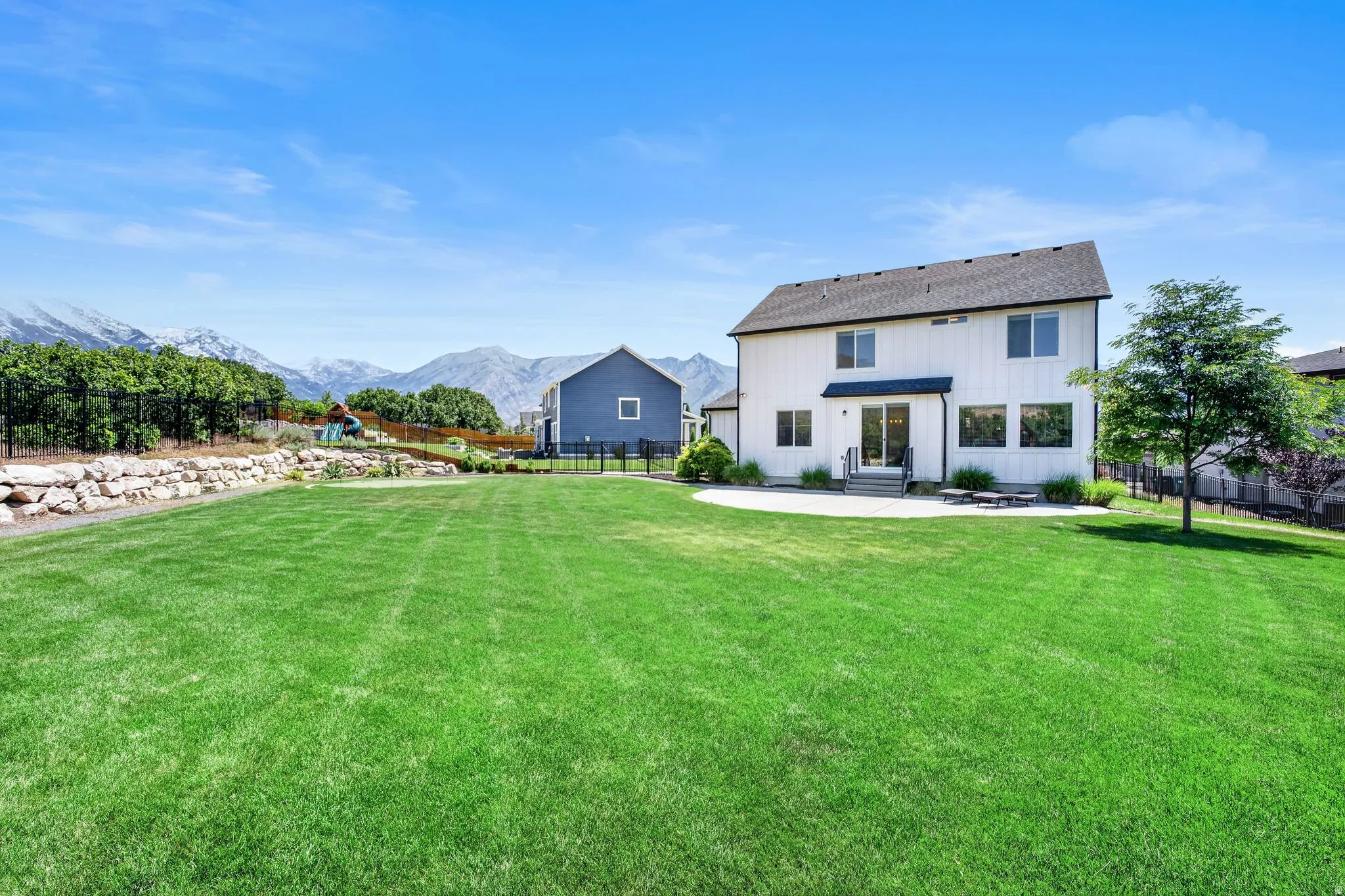 Expansive rear yard featuring a large manicured lawn, a concrete patio, and a retaining wall with rock landscaping