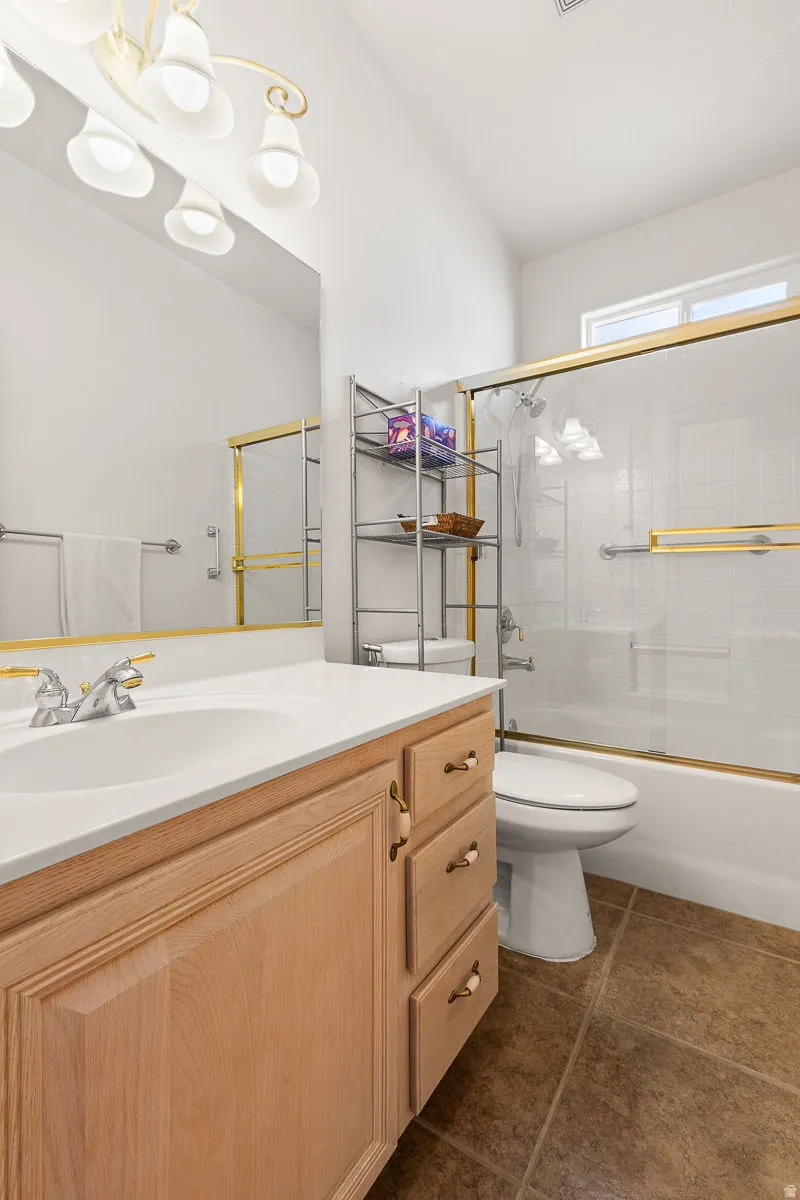 Bathroom featuring a light wood-finish vanity with a white countertop and integrated sink, gold-toned fixtures, a shower-tub combination with a sliding glass door, and brown tiled flooring