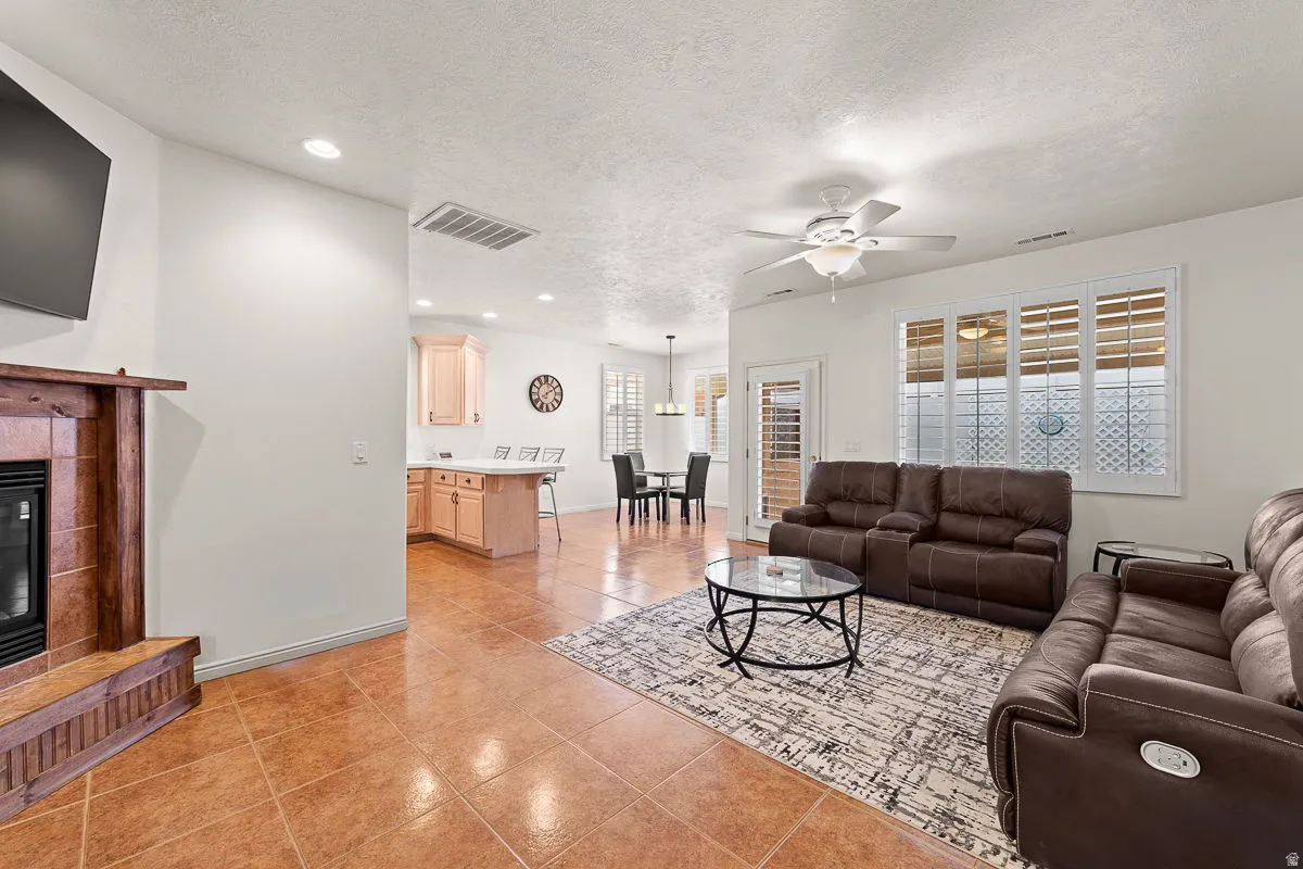 Open-concept living area featuring tile flooring, a fireplace with a wood mantle, white plantation shutters, and a ceiling fan