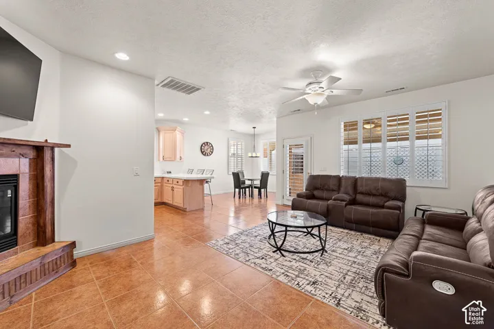 Open-concept living area featuring tile flooring, a fireplace with a wood mantle, white plantation shutters, and a ceiling fan