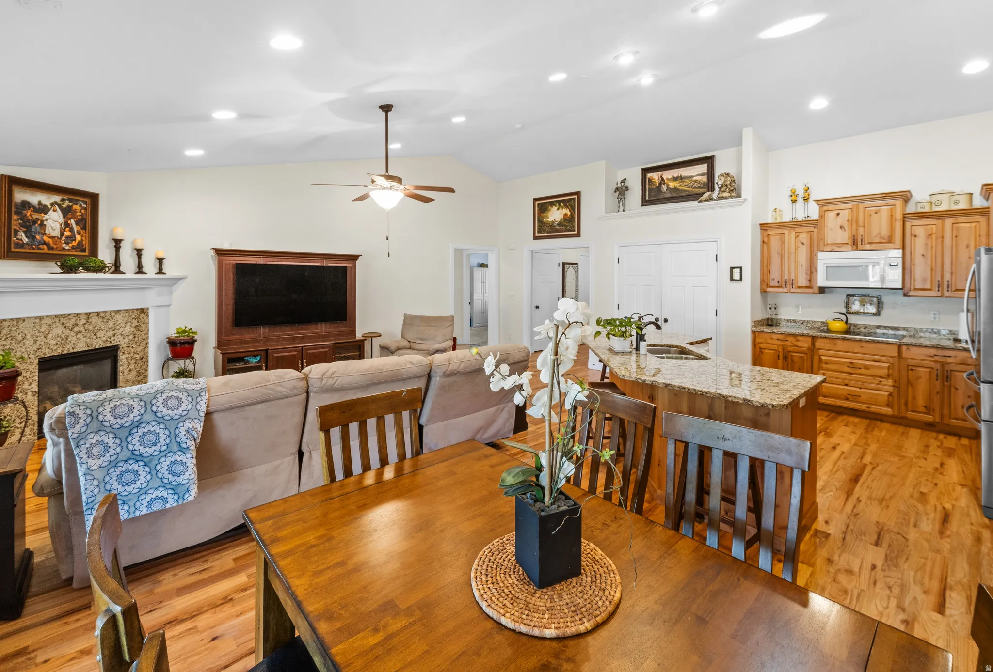 Open-concept living space with wood-finish flooring, a kitchen island with seating, natural wood cabinetry, a fireplace with a tiled surround, and vaulted ceilings with recessed lighting and a ceiling fan