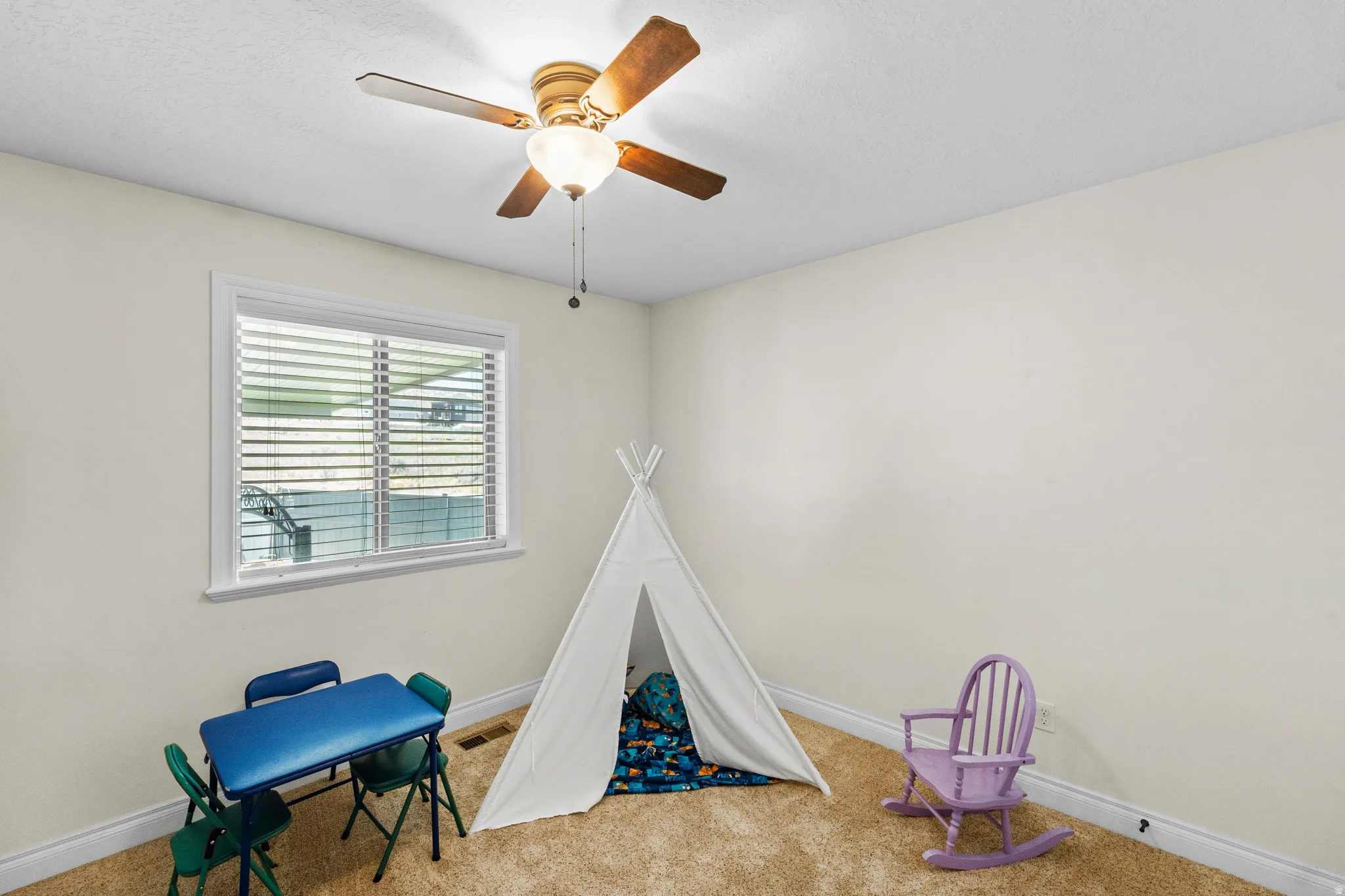 Room featuring neutral wall tones, textured carpet flooring, a white-framed window with horizontal blinds, and a ceiling fan with light fixture