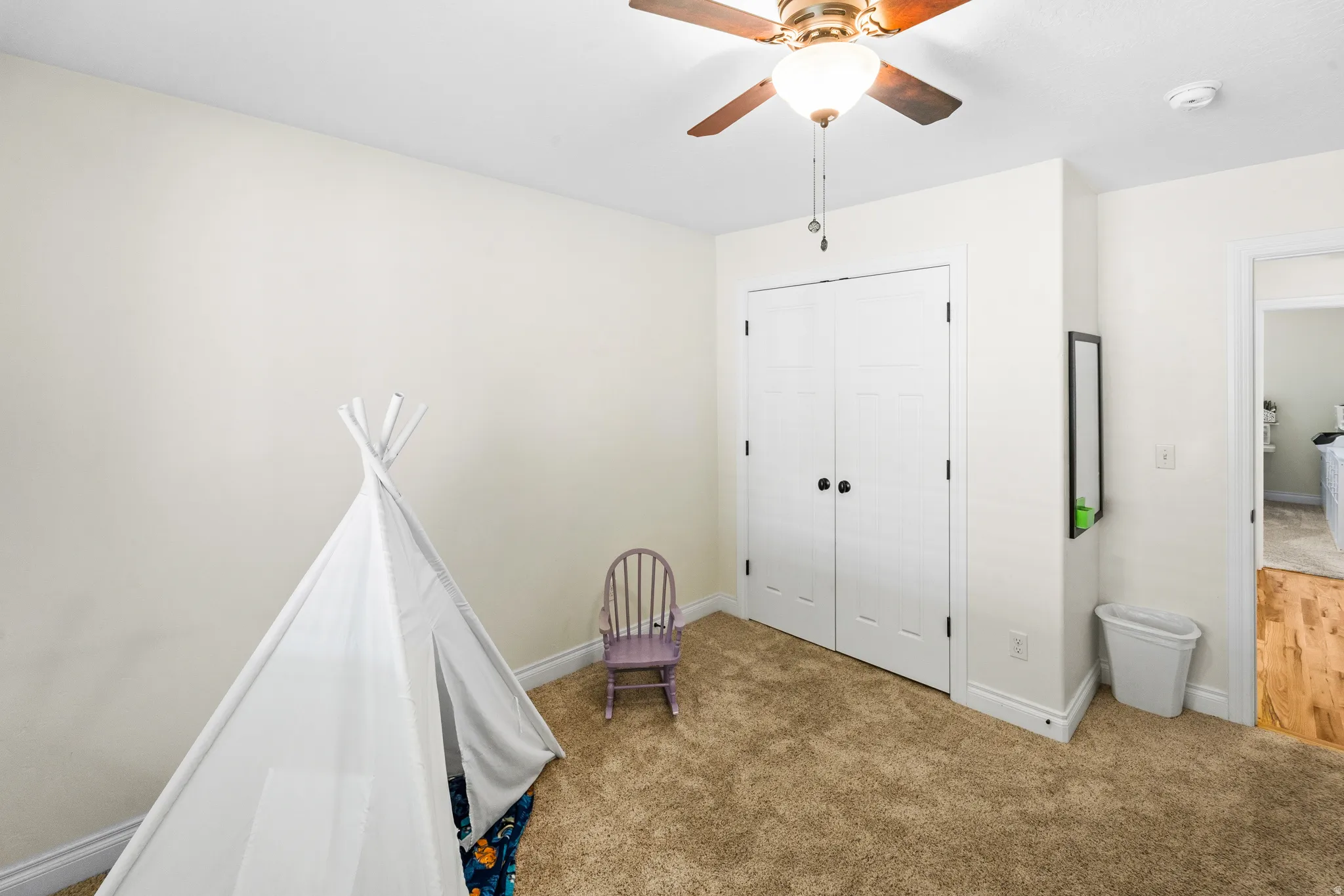 Carpeted room featuring a ceiling fan with integrated lighting, dual white bi-fold closet doors with black hardware, and an entryway with wood-finish flooring