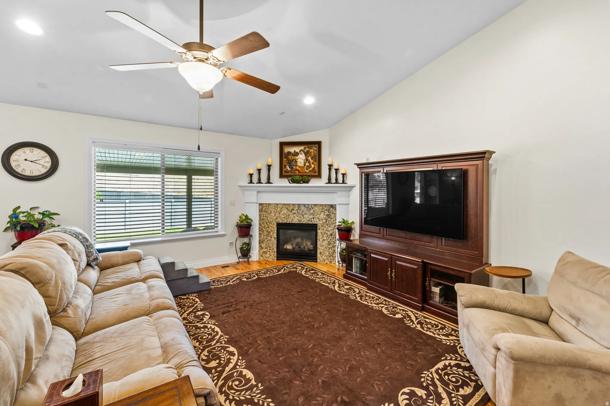 Living area featuring a vaulted ceiling with a ceiling fan, a fireplace with a tiled surround and white mantel, a large window with horizontal blinds, and wood-finish flooring