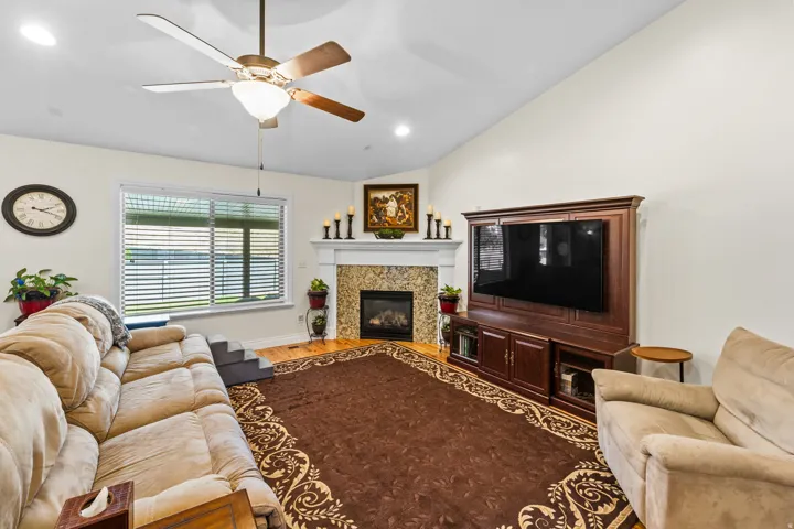 Living area featuring a vaulted ceiling with a ceiling fan, a fireplace with a tiled surround and white mantel, a large window with horizontal blinds, and wood-finish flooring