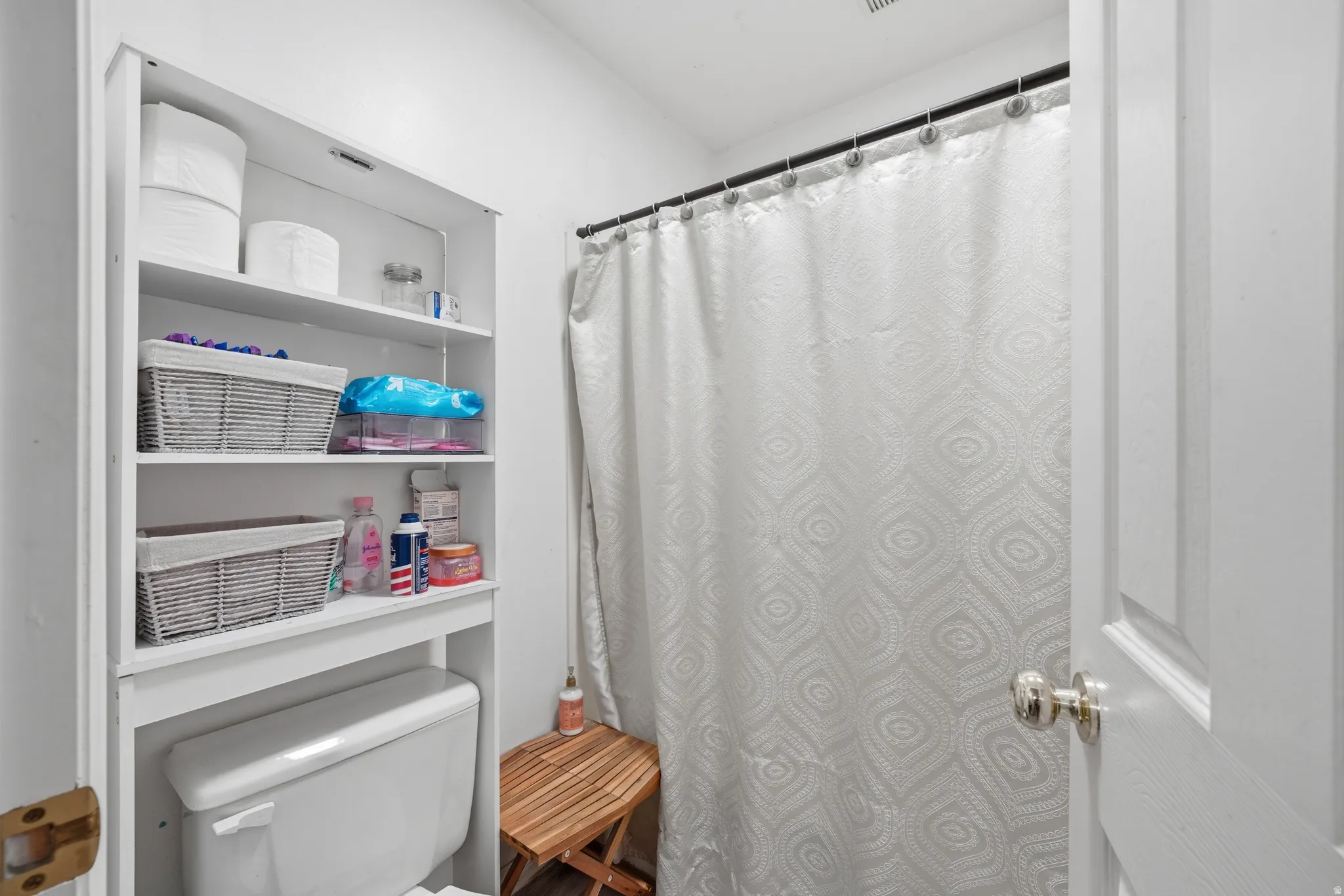 Compact bathroom featuring a white toilet with an integrated over-toilet shelving unit, a shower area with a black rod, and a paneled interior door with a polished chrome knob