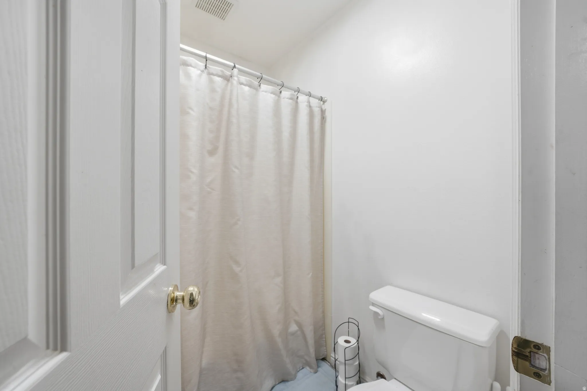 White paneled door with brass hardware opening into a bathroom featuring white walls, a standard toilet, and a shower/tub combination with a white curtain