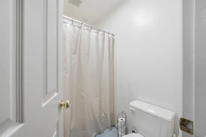 White paneled door with brass hardware opening into a bathroom featuring white walls, a standard toilet, and a shower/tub combination with a white curtain