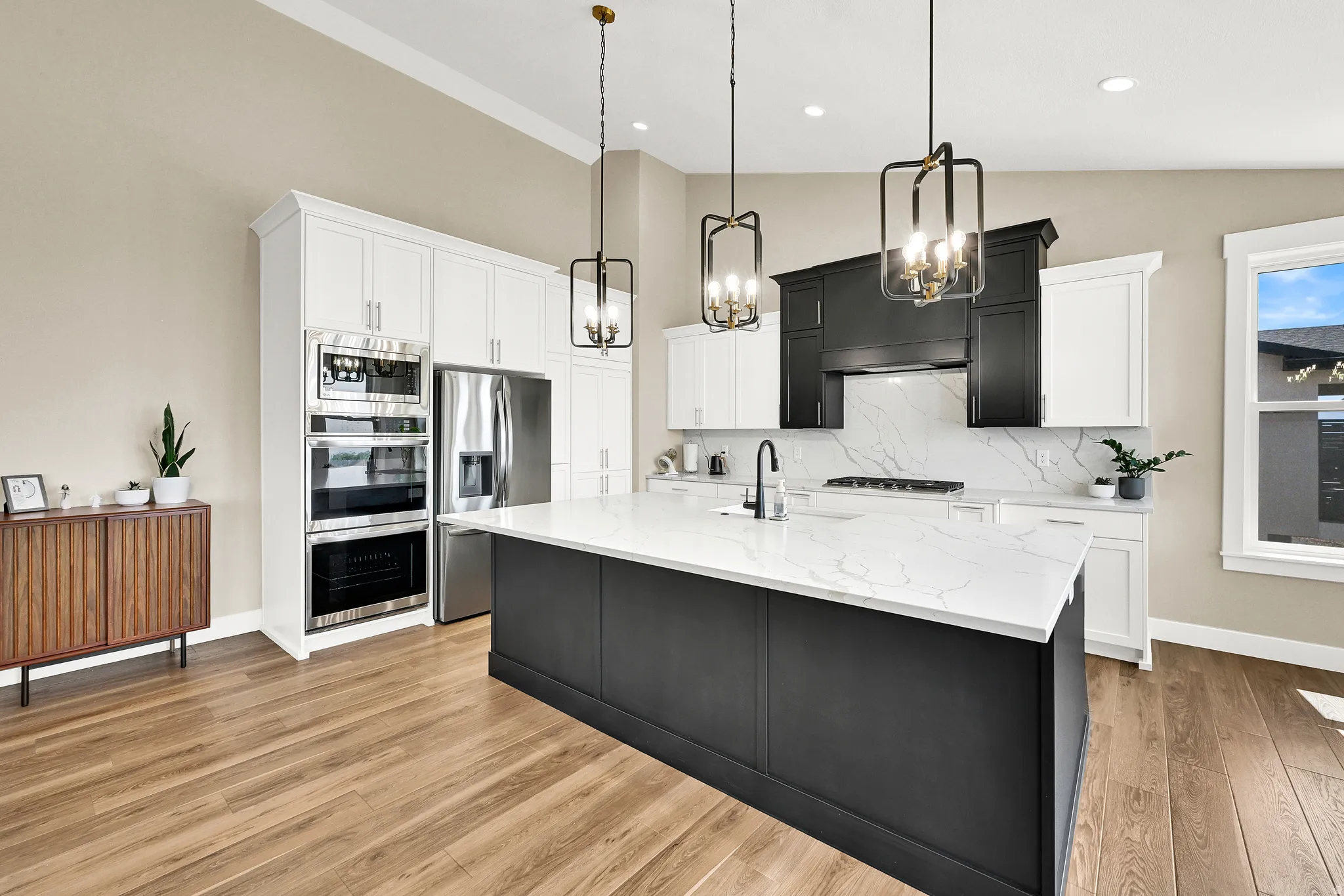 Spacious kitchen featuring a large island with a light-toned countertop, stainless steel appliances, wood-finish flooring, white perimeter cabinetry, and black accent cabinetry with a matching hood