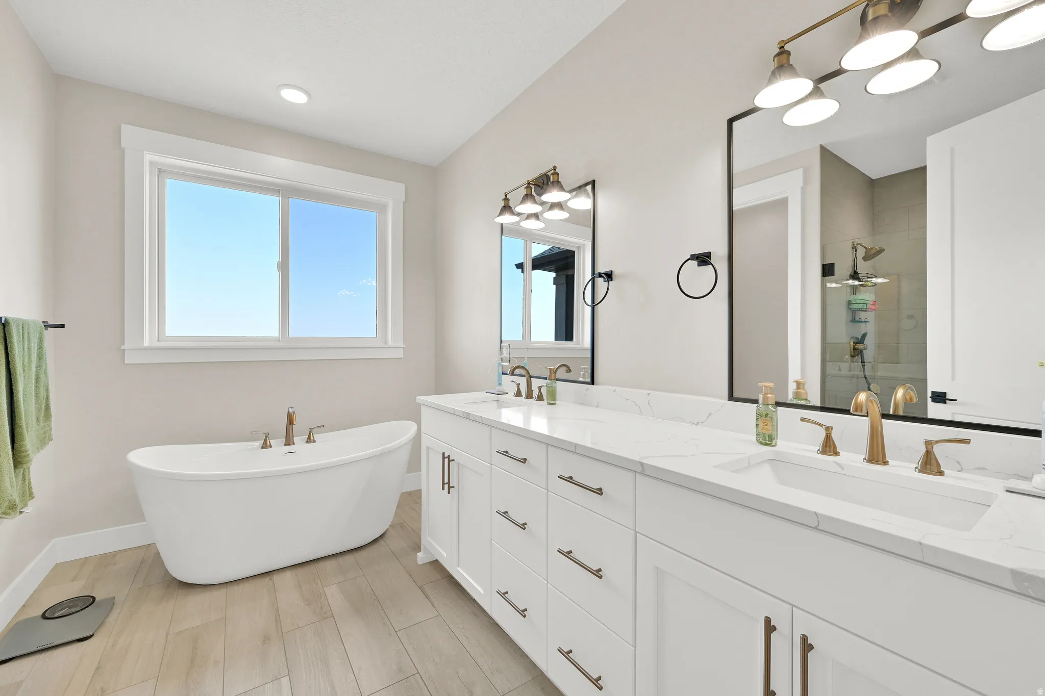 Spacious bathroom featuring a freestanding soaking tub, dual vanity with white cabinetry, quartz-finish countertops, and wood-finish flooring