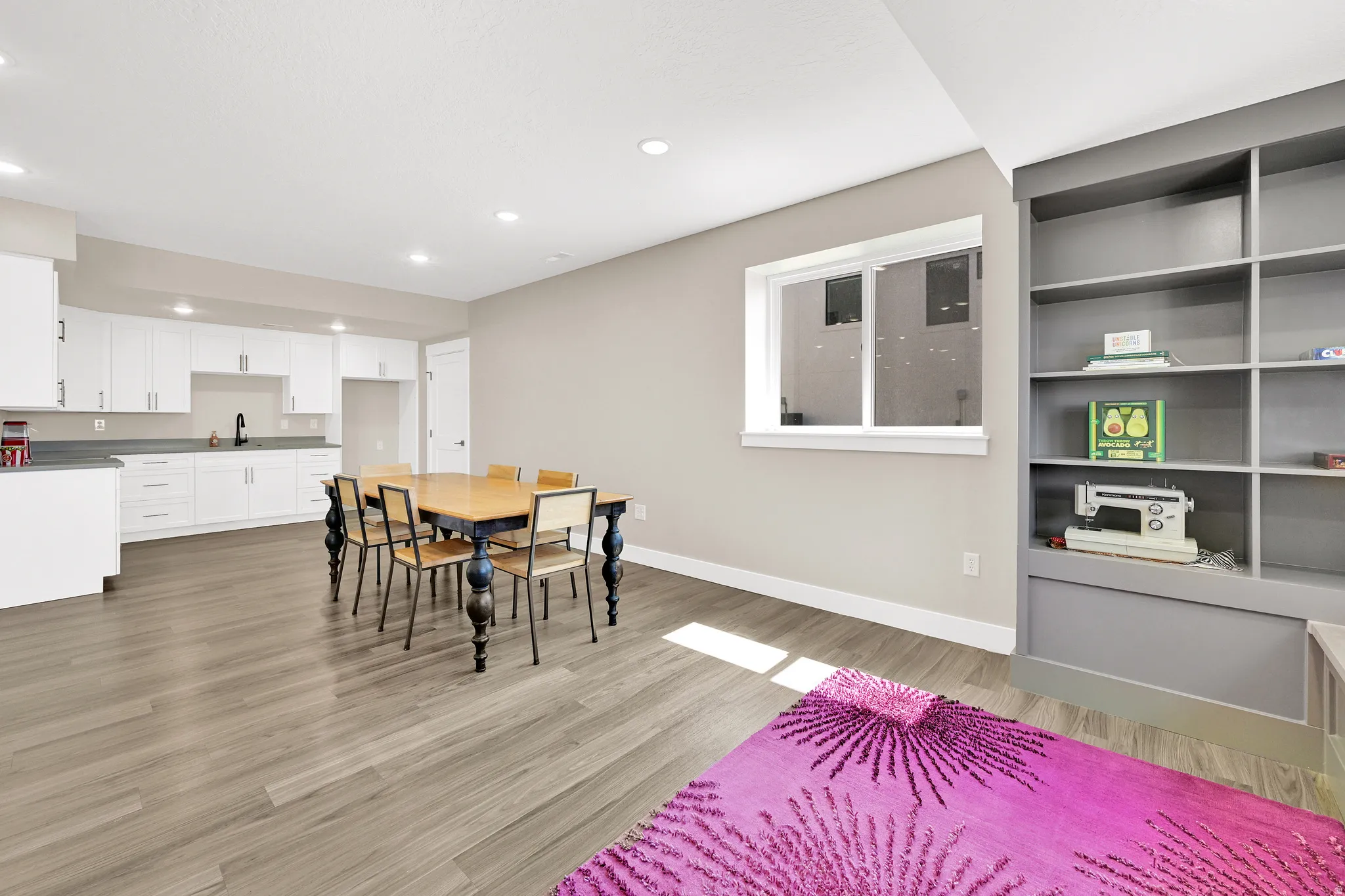 Spacious open-concept room featuring light wood-finish flooring, a full kitchen with white cabinetry and dark countertops, and a built-in shelving unit