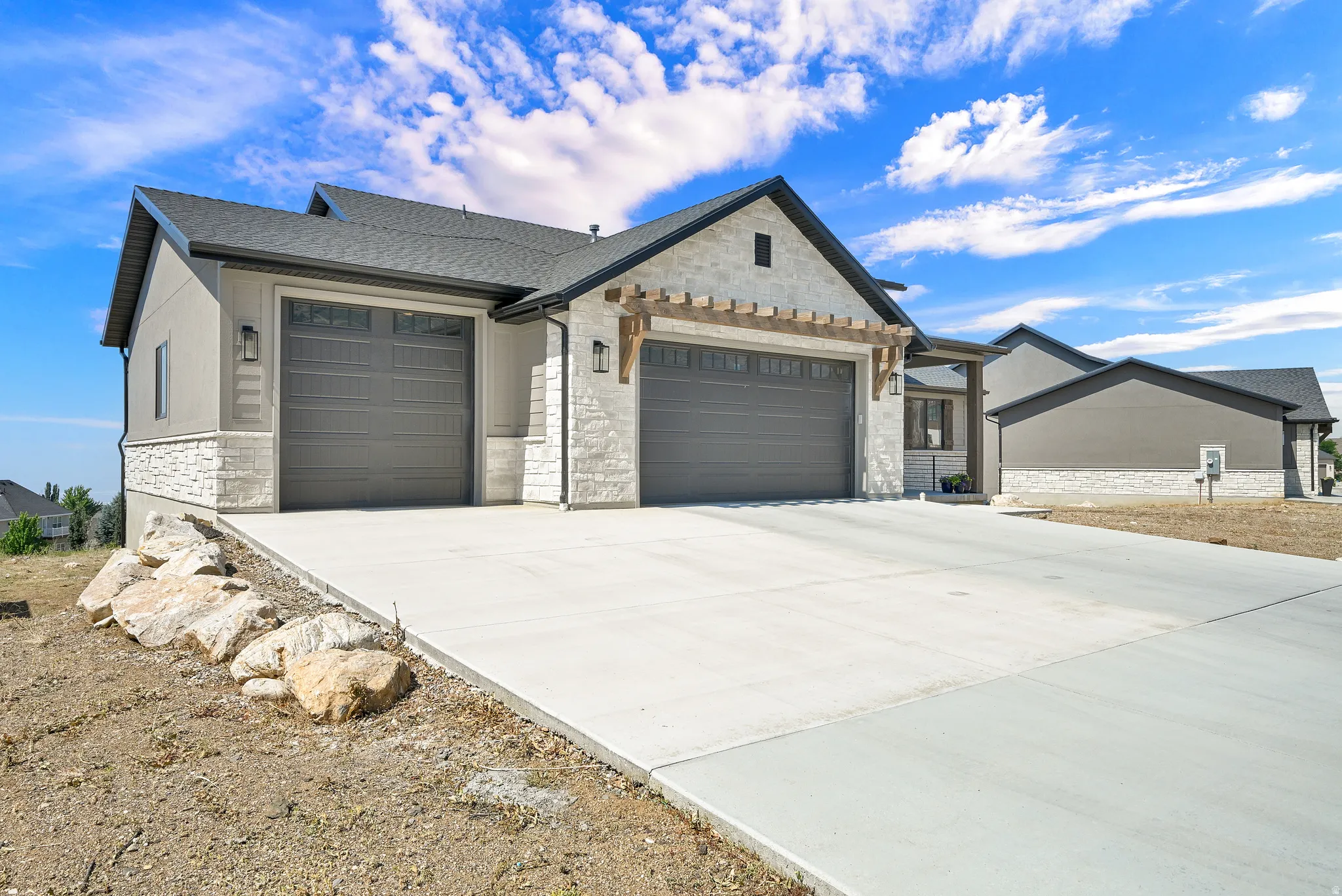 Contemporary exterior featuring stucco and stone accents, a multi-car driveway, two garages with carriage-style doors, and decorative wooden pergola details over the front garage