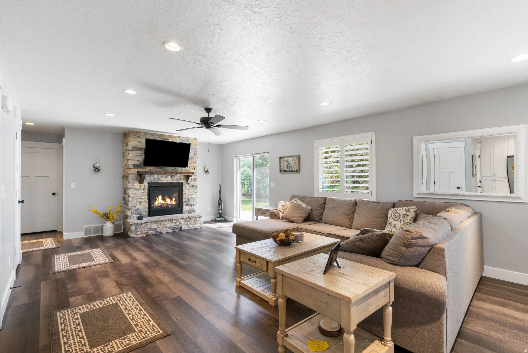 Living area featuring wood-finish flooring, a stone-clad fireplace with a wood mantel, a ceiling fan, recessed lighting, and white window shutters