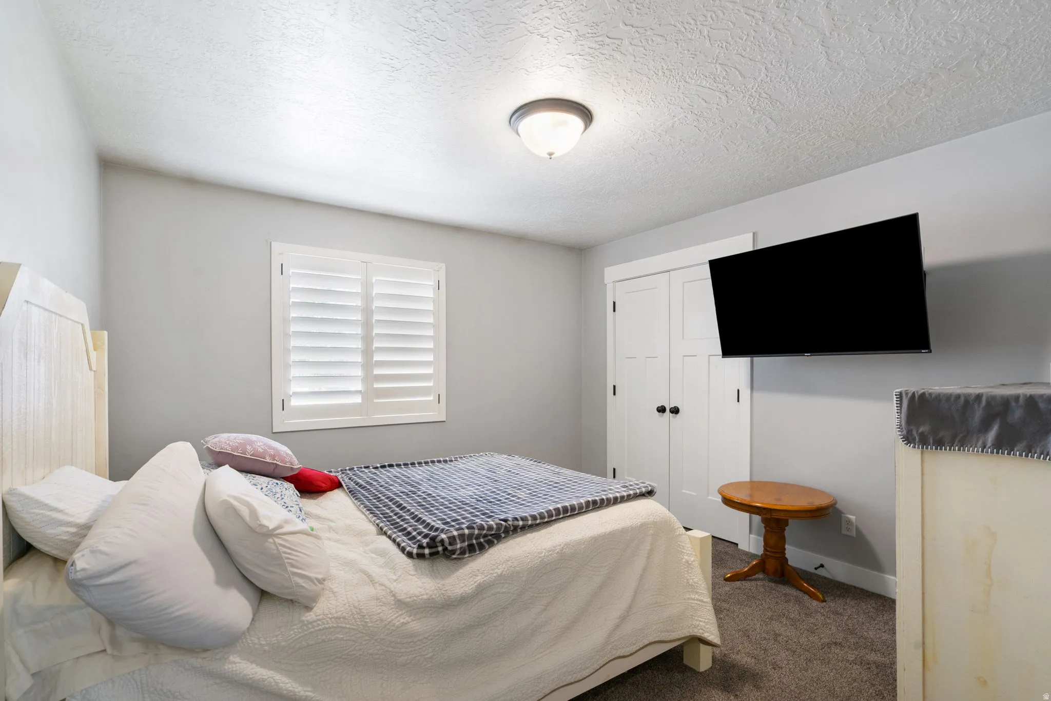 Bedroom featuring a window with white plantation shutters, neutral gray wall color, textured carpet flooring, and a ceiling-mounted light fixture