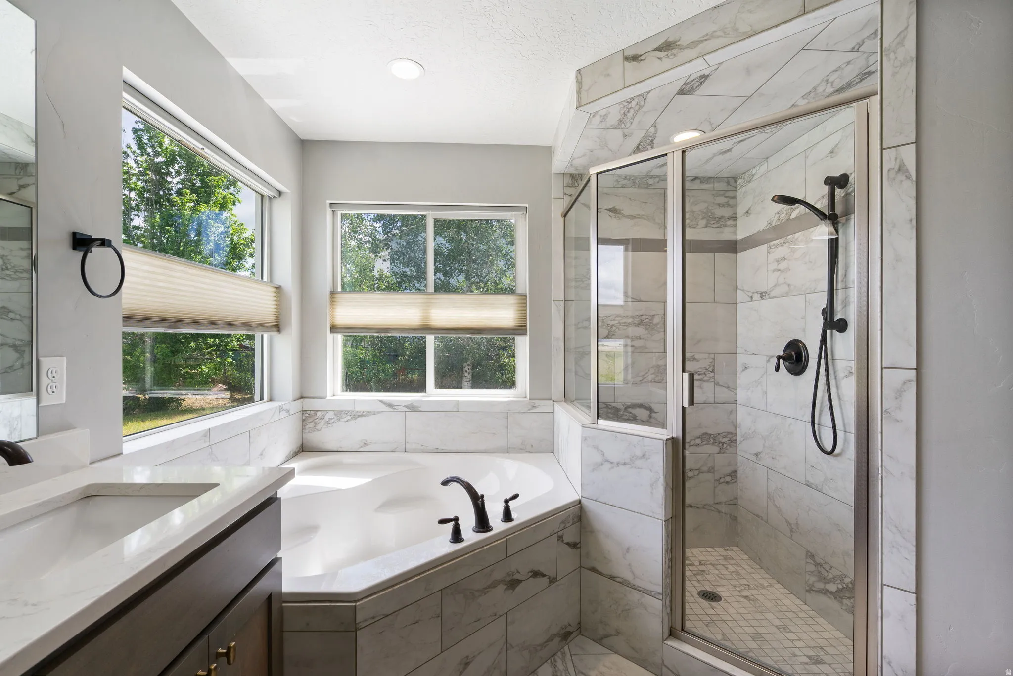 Bathroom featuring a corner soaking tub with matte black fixtures, a walk-in shower with glass enclosure and mosaic tile floor, and a double vanity with white countertops