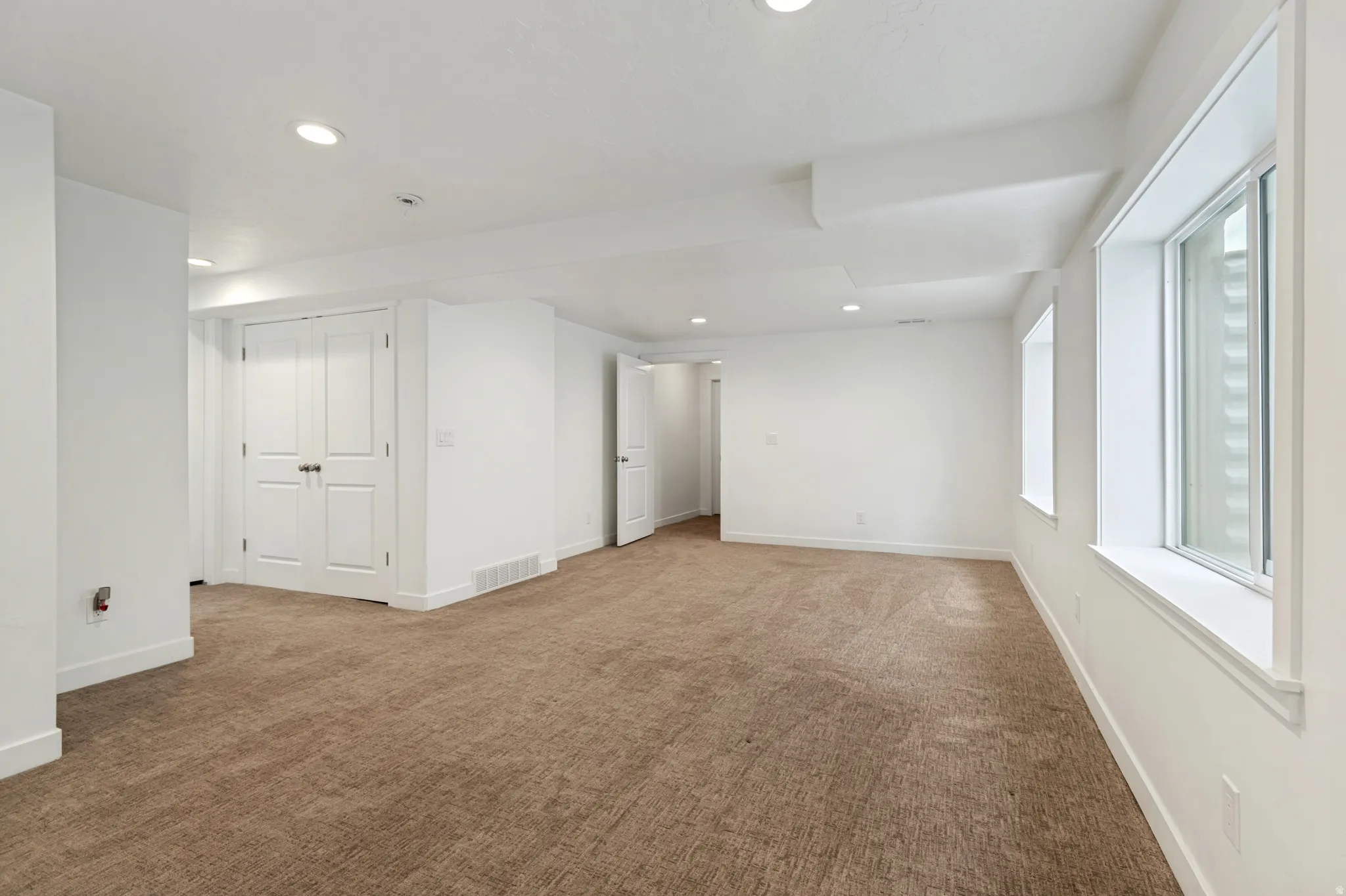 Spacious room with light brown carpet flooring, multiple windows, recessed lighting, white walls, and white bi-fold doors