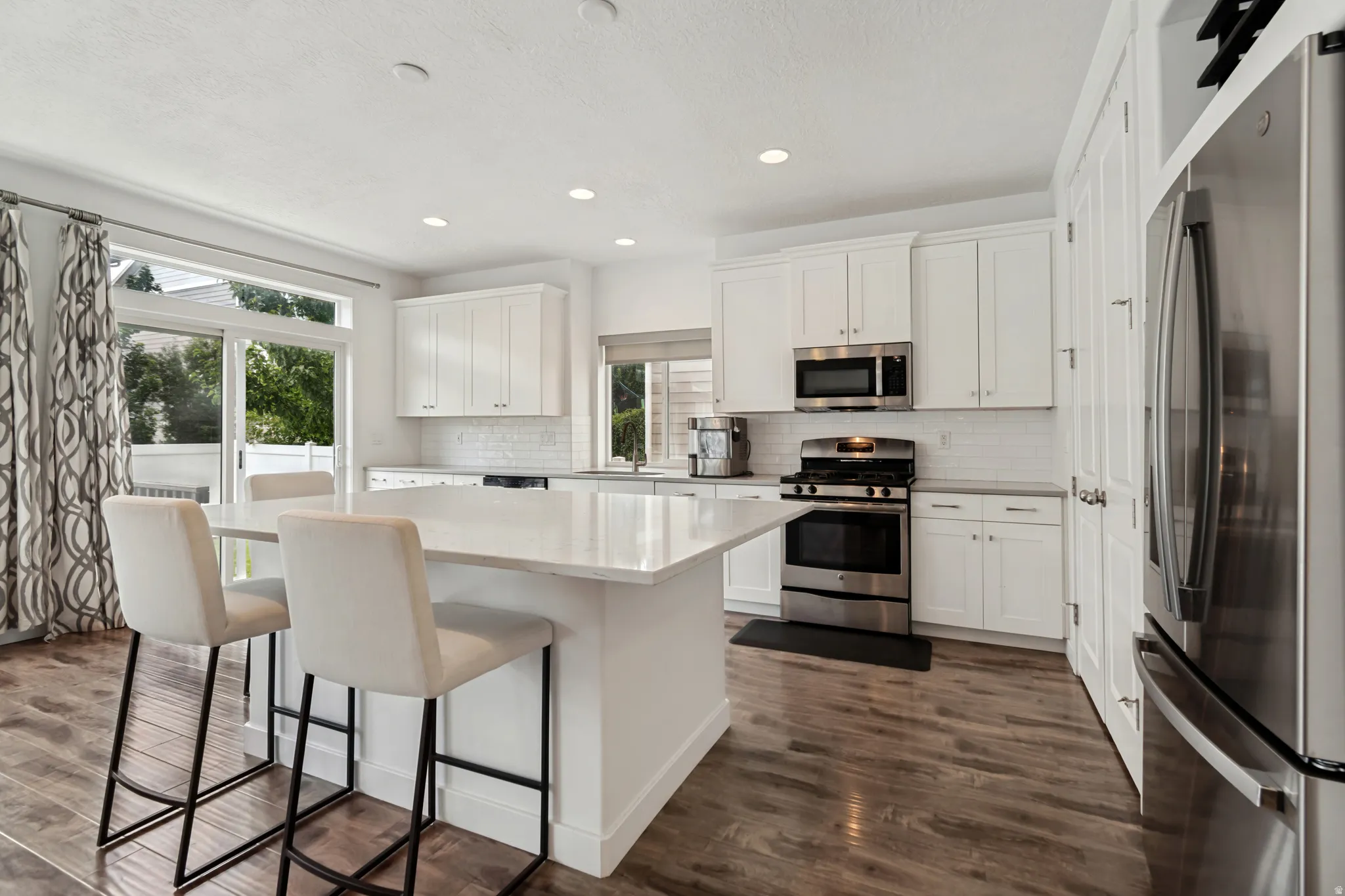 Contemporary kitchen featuring a large center island with a stone countertop, white shaker-style cabinetry, stainless steel appliances, wood-finish flooring, and a subway tile backsplash