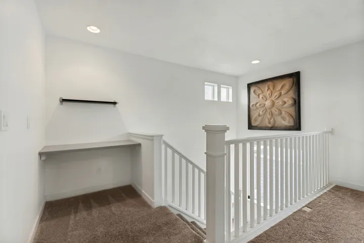 Second-story landing featuring a built-in desk area with a wall-mounted shelf, recessed lighting, white balusters, and a neutral carpet