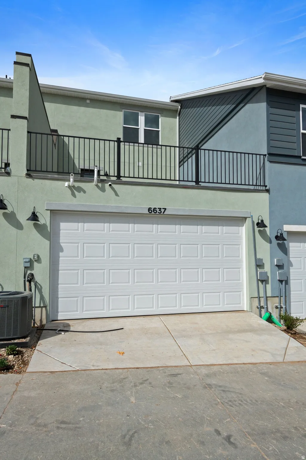 Contemporary exterior featuring light green and blue siding, a two-car garage with a panel door, and an upper-level balcony with black metal railing