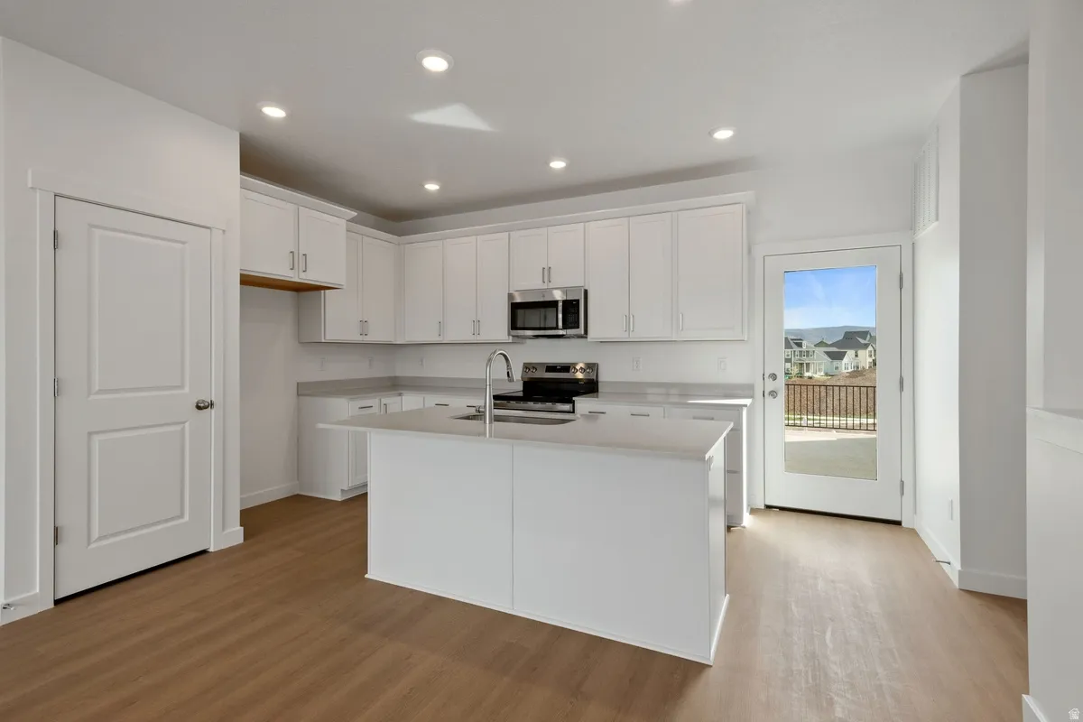 Kitchen featuring white cabinetry, light-toned countertops, a central island with an undermount sink, stainless steel appliances, and wood-finish flooring