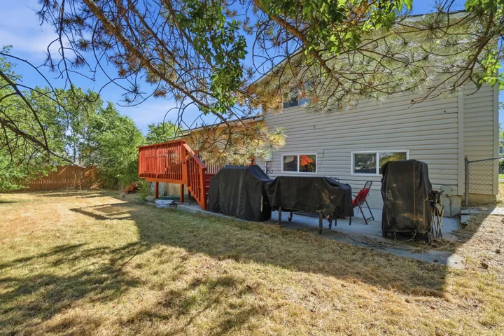 Rear exterior featuring light grey siding, a red-stained wood deck with stairs, a concrete patio, and a chain-link fence