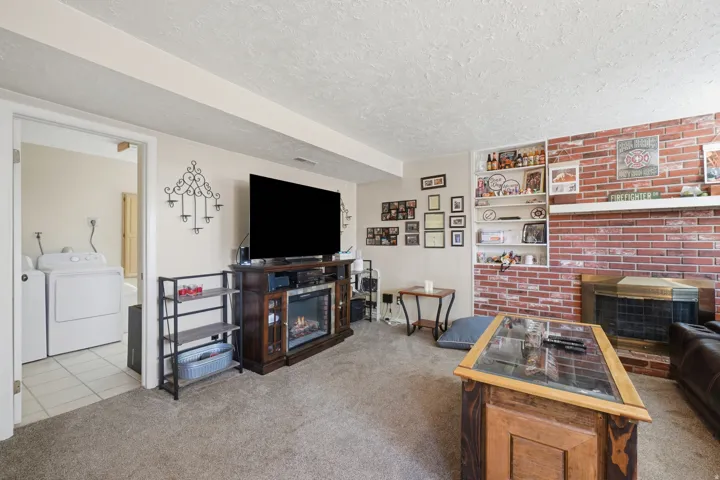 Carpeted living area featuring a brick accent wall with a wood-burning fireplace, built-in shelving, and a doorway to a tiled laundry area