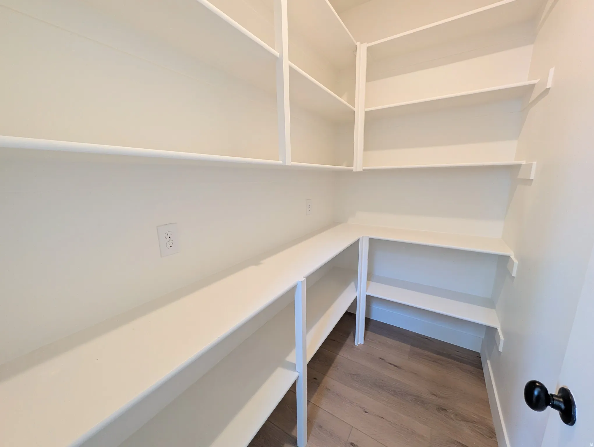 Walk-in pantry featuring extensive white shelving, wood-finish flooring, and an electrical outlet