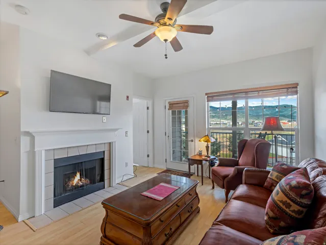 Living area featuring a fireplace with a tiled surround and white mantel, wood-finish flooring, a ceiling fan with integrated lighting, and a glass-paneled door opening to an outdoor space