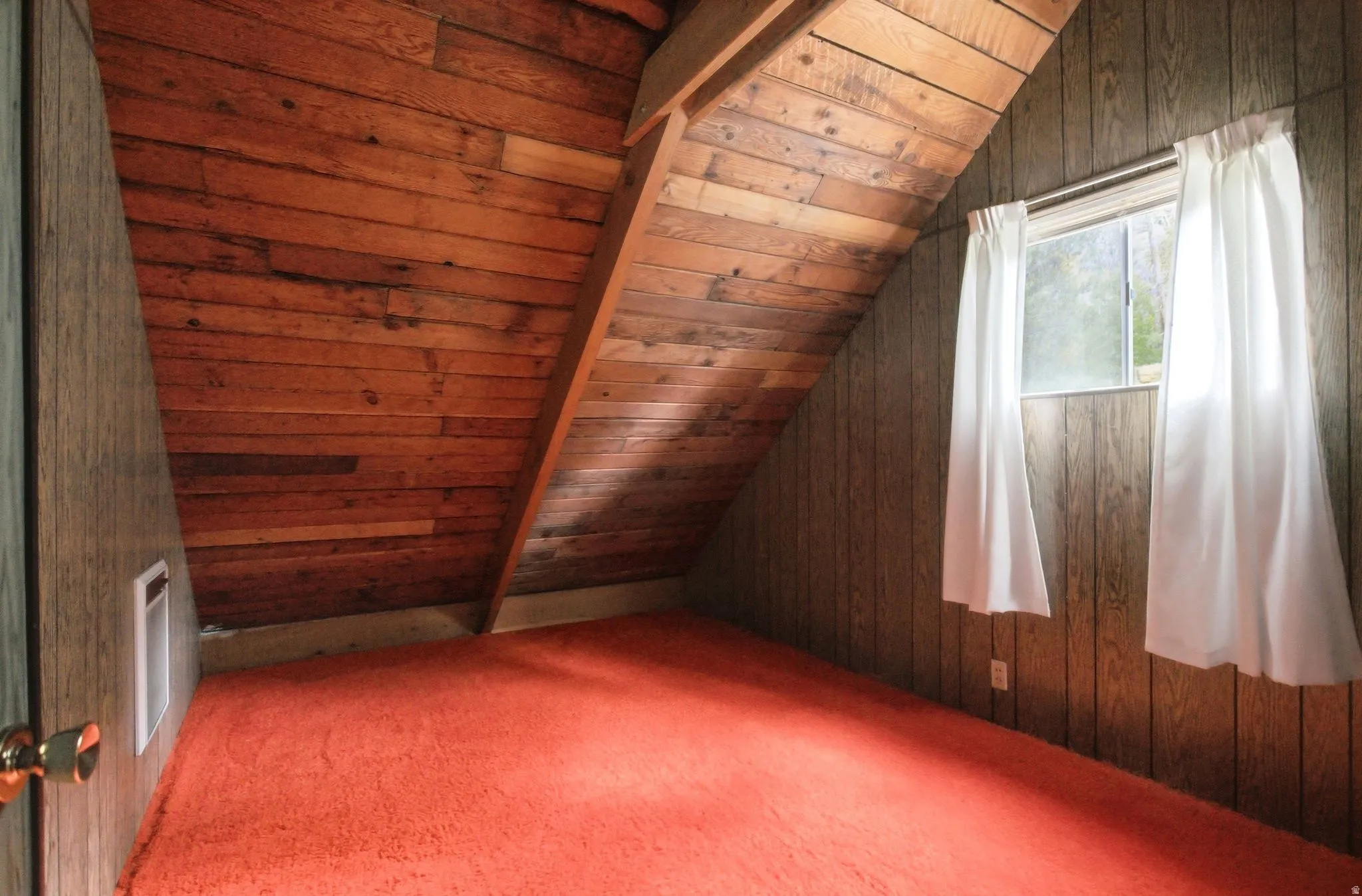 Attic room featuring sloped wood-paneled ceilings and walls, orange carpeting, and a single window with white curtains
